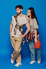 couple posing in street wear on blue backdrop, woman with bold makeup, bearded man, baseball cap