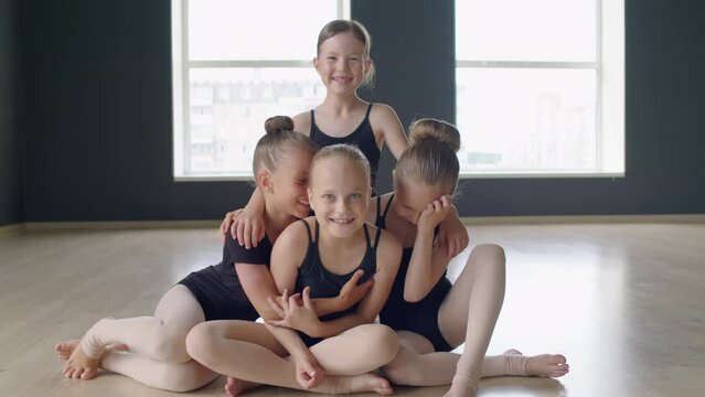 Portrait of positive Caucasian small gymnasts in leotards hugging each other while sitting on floor and looking at cameraPortrait of positive Caucasian small gymnasts in leotards hugging each other wh