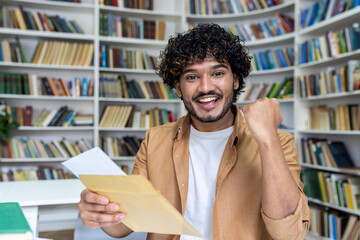 Portrait of young successful student, man received letter of notification of good results of interview exam, hispanic man joyfully looking at camera inside library, holding mail envelope.