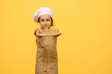 little girl in chef hat and apron, smiles and holds out at camera a wooden board, isolated over yellow studio background