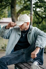 Young adult male is seated on a bench in a park, tying his baseball cap.