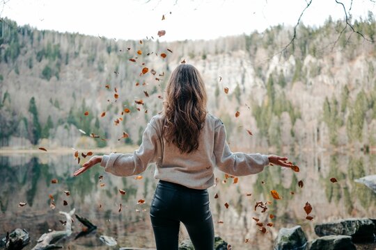 Young woman wearing tight jeans and a sweater, standing in front of a lake, throwing leaves.