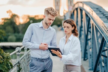 businesswomen and businessman standing on a bridge during sunset, discussing something on the tablet
