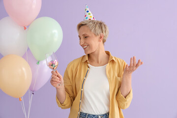 Mature woman with glass of confetti and balloons on lilac background