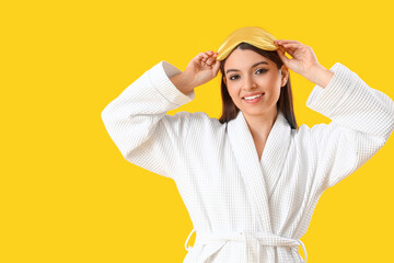 Young woman in gown with sleeping mask on yellow background