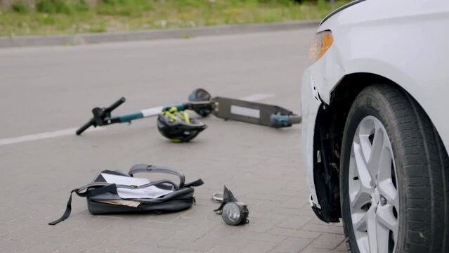 A road accident between a luxury white car and an electric scooter on an asphalt road. The cyclist's helmet, backpack and pieces of the car are thrown to the ground. The concept of accidents