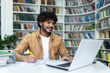 Indian student with curly hair studying sitting among books on shelves, man watching video course writing in notebook smiling contentedly.