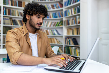 Young hispanic student studying inside university campus academic library among bookshelves, man smiling and using laptop.