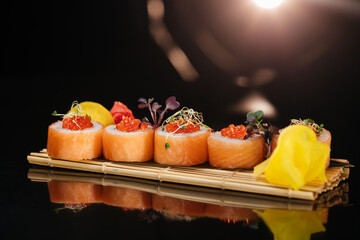 Four big rolls of Philadelphia Special sushi with salmon, cream cheese, red caviar, yellow and red ginger, arranged on a board made of wooden sticks tied with string, on a black background