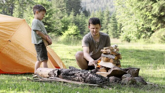 Dad and son sit together by a bundle of firewood and light a fire near the tent. The spirit of adventure and adventure. Hiking and camping. Travel concept 4K.