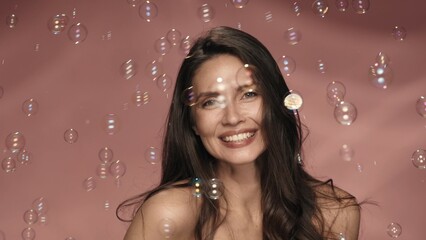 Joyful, playful woman surrounded by soap bubbles. A seminude woman with flowing hair in the studio on a pink background. Lightness, childhood. - obrazy, fototapety, plakaty