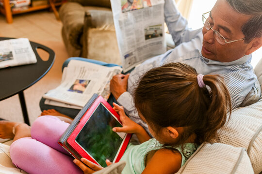 Grandfather holding newspaper looking at granddaughter's tablet