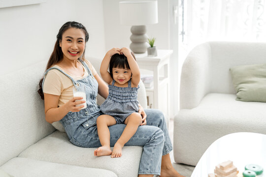 Little asian girl and her mom drinking milk sitting on sofa