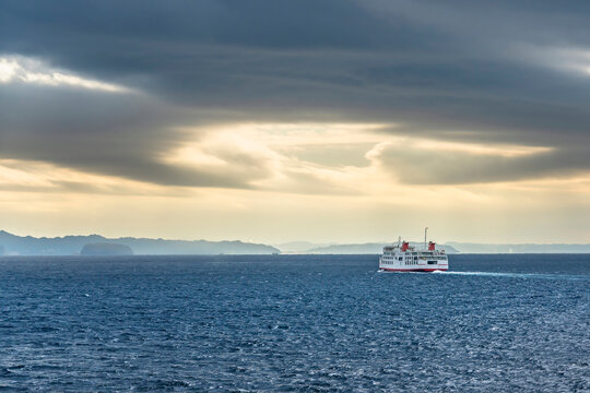 Ferry ship on the sea of the Uraga channel sailing towards the Kanaya port along the coasts of the Boso peninsula in the morning light below a stormy sky.