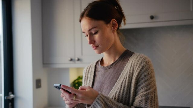 Young woman taking a break with cup of coffee standing by kitchen counter checking social media or message on mobile phone - shot in slow motion