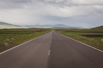 Straight tarmac road through green grasslands of Javakheti Plateau with white car driving in the distance and Caucasus Mountains in the background, Georgia.