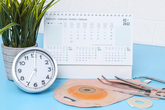 Clock with time seven, calendar for february 2022, surgical instruments, stethoscope, colostomy bag on a white and blue.