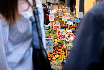 Women buying fruit and vegetables in a street market in Naples, Italy. Women seen from behind, signs with prices, oranges, bananas and seasonal fruit.