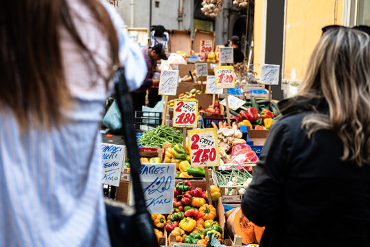 Women buying fruit and vegetables in a street market in Naples, Italy. Women seen from behind, signs with prices, oranges, bananas and seasonal fruit.
