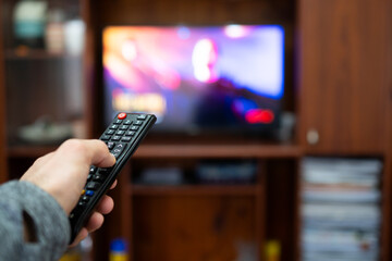 Hand of a person in the foreground holding a remote control and pointing to a TV in the background...