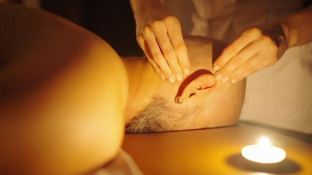 Ear massage in the SPA salon. Slow motion, candlelight, bottom angle. Women's hands massage a man.