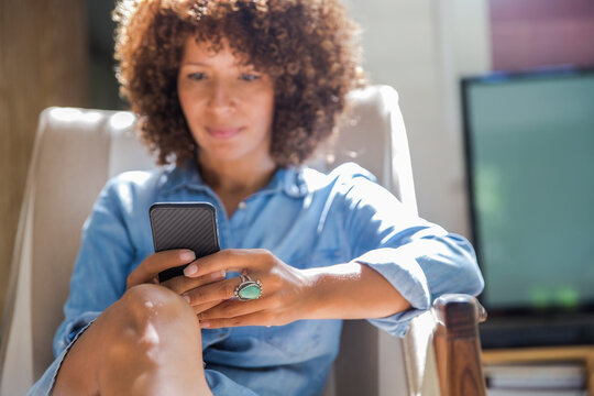 Relaxed Woman With Curly Hair Using Mobile Phone On Armchair At Home