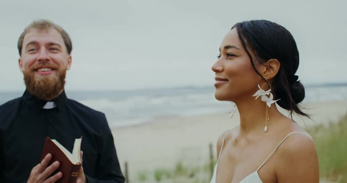 The bride and groom laugh standing in front of the priest on the seashore