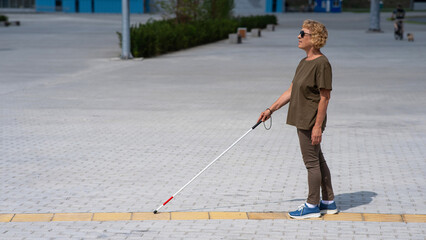An elderly blind woman walks with a cane along a tactile tile. 