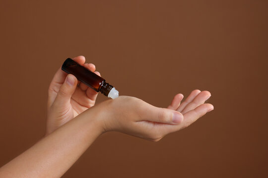 Woman with roller bottle applying essential oil onto wrist on brown background, closeup