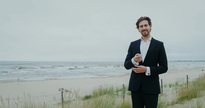 The groom smiles looking at the camera and adjusting cufflinks on his sleeve while standing on the seashore