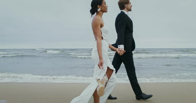 The bride and groom hold hands walking on the beach, the bride holds shoes in her hands