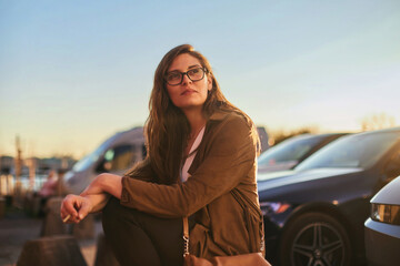 A young woman is seated in a parking space, smoking at sunset.