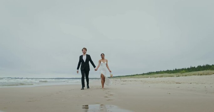 The bride and groom hold hands walking on the beach, the bride holds shoes in her hands
