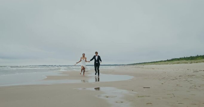 The bride and groom holding hands ran along the sandy beach, the bride holds her shoes in her hands