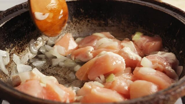 Pieces of chicken fried in a cast-iron skillet in oil are mixed with a wooden spatula. Close-up