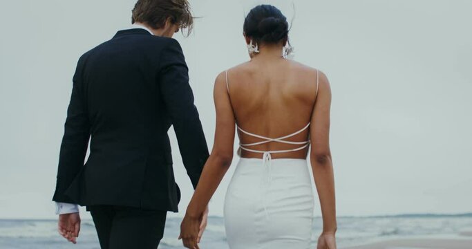 The bride and groom hold hands and smile at each other while walking on the beach
