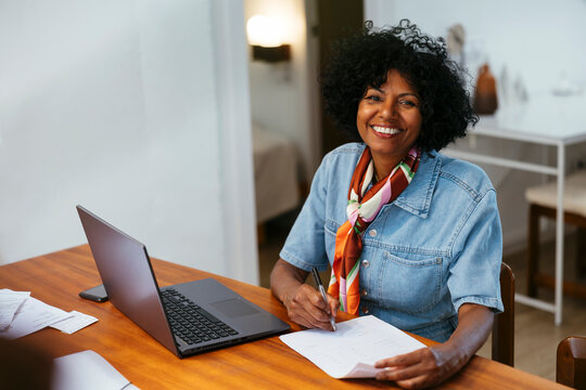 Cheerful woman looking at camera with notebook and laptop at home