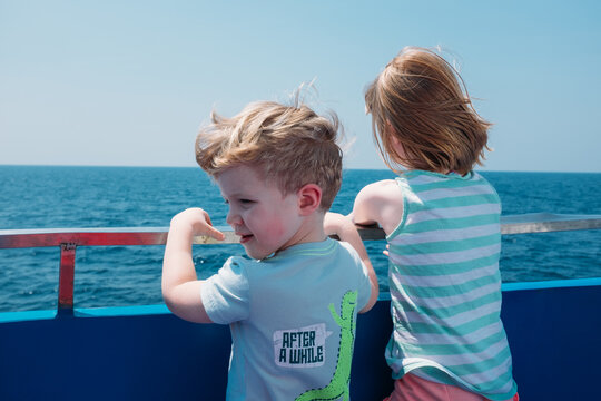 Two Young Kids Look Over The Side Of A Boat At Sea