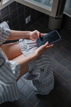 Faceless woman sitting on a toilet and scrolling her phone