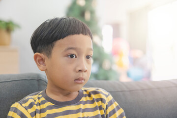 Close-up Asian boy with black hair wearing a yellow-gray striped t-shirt looking at the TV in the background of christmas trees and wooden furniture. Child makes funny faces in his house happily.