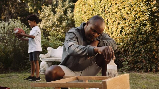 Black father focusing on DIY project at home in the garden while son plays with football