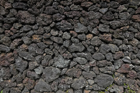 A wall made up of dark, volcanic pumice rocks is shown in a closeup, exterior view during the day.