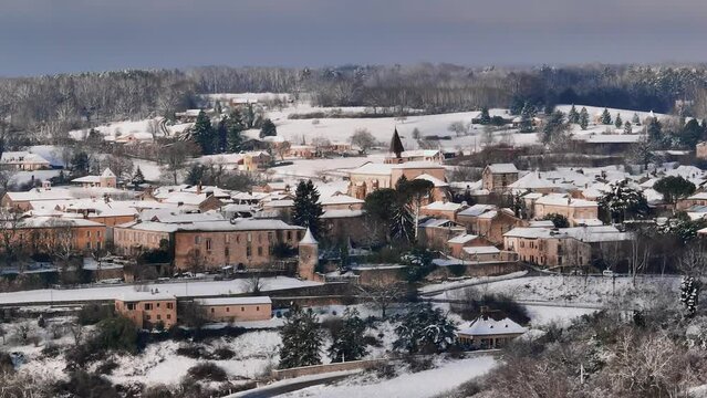 Small village under the snow in France, Monpazier in the Dordogne, morning rises over the countryside, circular drone shot around the church illuminated by the morning sun.
