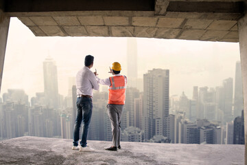 Back view of two Indian male engineer and architect wearing helmet and vest discussing project at construction site with lots of buildings in the background. Wide angle, Full length shot.