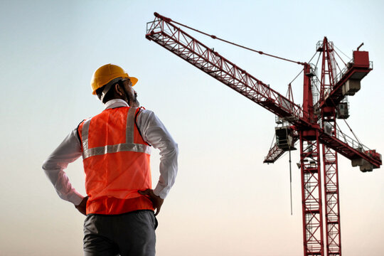 Back view of Indian male engineer wearing helmet and vest standing and supervising progress of construction project with crane in the background, Copy space. Low angle shot.