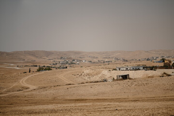 In a remote area of Israel's Negev Desert, a rural Bedouin village is located amid barren hills. Houses, barns, and other small structures fill the scene. The background is a sunny, hazy sky.