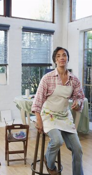 Vertical video of portrait of happy biracial woman in apron holding brushes in studio, slow motion