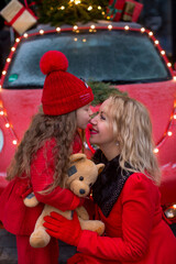 Six-year-old daughter with a teddy bear kisses her mother on the nose in front of a red car with Christmas lights and gift boxes