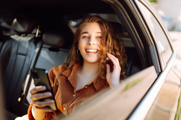 Cheerful woman with curly hair and stylish clothes sits in the back seat of a car, uses a mobile phone. The concept of business, technology, travel, online communication.