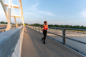 As the sun sets, she runs across the bridge, a bottle in her hand, periodically glancing at her watch. The vibrant hues of the sky mirror her determination
